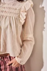 Blusa de color arena con diseño romántico, presenta volados en los hombros, detalles de encaje en el pecho y mangas largas con puños elásticos.