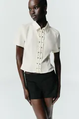 Camisa de manga corta en tejido popelín color crudo, con cuello solapa y cierre frontal con cremallera oculta. Presenta detalles de volantes verticales y tachas metálicas en la parte delantera.