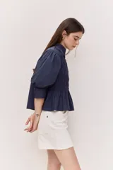 Camisa azul marino de cuello clásico, mangas abullonadas a tres cuartos y detalle fruncido en el frente. Presenta una silueta amplia y diseño corto con vuelo en el ruedo.