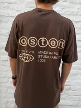 Remera marrón de manga corta, corte oversize, con un pequeño logo de un globo terráqueo estilizado y el texto "STUDIO ARCHIVE" estampado en el pecho.