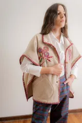 Poncho de paño color beige con flores bordadas en el frente y detalles de costura artesanal en los bordes en color bordó. Cuenta con bolsillos laterales y cierre frontal con botones de madera.