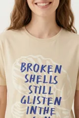 Remera de manga corta color beige, confeccionada en 100% algodón, con cuello redondo y estampa frontal con texto en tonos rosados.