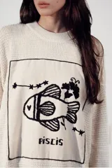 Sweater de tejido de punto color crudo, con cuello redondo y diseño oversize. Presenta un parche frontal bordado a mano en hilo negro con la temática del signo zodiacal Piscis, que incluye un pez, estrellas y la inscripción 'Piscis'.