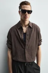Camisa de corte relajado confeccionada en tejido ligero de algodón con textura de cuadros. Presenta cuello solapa, manga larga con puños abotonados y cierre frontal de botones.