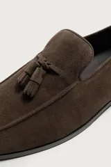 Mocasines de vestir para hombre confeccionados en piel con acabado en gamuza de color marrón, con detalle de borlas en el empeine y suela plana con ligero tacón.