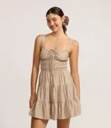 Vestido corto color beige, con escote recto, breteles medianos y lazo decorativo en el centro del busto. Presenta costura fruncida en la cintura y falda con babados en capas.