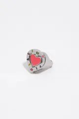 Anillo de metal plateado con diseño de corazón. Presenta un centro esmaltado en color rosado y un borde con letras de colores y detalles grabados.