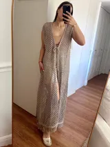 Chaleco largo sin mangas, tejido en punto calado tipo red con hilo de algodón y lurex, color marrón. Presenta escote profundo en V, tiras para atar en el frente, tajo trasero y flecos en el ruedo.