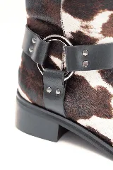 Bota corta de cuero con pelo de vaca en diseño animal print de manchas marrones y blancas, con punta cuadrada, taco bajo y correa de cuero negro con tachas y una argolla metálica alrededor del tobillo.