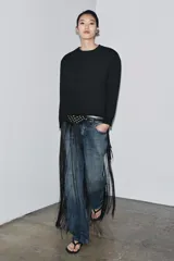 Cinturón tipo fajín negro de 10 cm de ancho, confeccionado en piel 100%. Presenta una sección superior con tachas doradas y ojales metálicos, y la parte inferior terminada en flecos largos. Se ajusta al frente con una lazada de cordón negro.