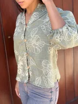 Camisa de corte holgado en color verde con estampado floral lineal en tono crema. Presenta cuello mao, cierre frontal con botones y mangas tres cuartos con puños elásticos.