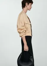 Chaqueta tipo bomber color beige, con cierre frontal de cremallera, cuello redondo, bolsillos de solapa en el pecho y cintura y puños elásticos.