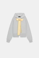 Chaqueta de punto con capucha, manga larga y cierre frontal con cremallera. Presenta un diseño de lazo decorativo en color amarillo en la zona del cuello.