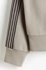 Campera deportiva de color gris topo, con cuello alto, cierre frontal bidireccional y bolsillos laterales. Presenta franjas laterales negras en las mangas y puños y bajo de punto acanalado.