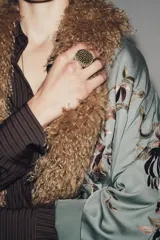 Maxi anillo metálico de forma circular con un engaste central lleno de pequeñas piedras negras brillantes. El borde exterior del anillo presenta un diseño labrado con textura.