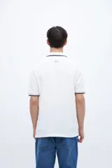 Remera polo blanca con cuello y puños acanalados con rayas azules y rojas. Presenta una media cartera de botones y un bordado en el pecho con la palabra "Uniform" en azul.