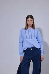 Camisa blanca de algodón con pechera decorada con alforzas y puntillas. Presenta cierre frontal con botones, mangas tres cuartos con frunces y detalles de puntilla, y un diseño de corte holgado con ruedo redondeado.