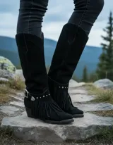 Bota de caña alta estilo western confeccionada en cuero vacuno negro, con flecos decorativos, estribo metálico con hebilla y tachas. Presenta taco de madera y punta fina.