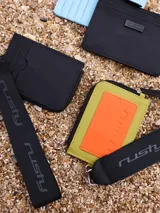 Tarjetero Rusty Runoi con cierre y correa para colgar, confeccionado en poliuretano color verde con aplique naranja y logo de la marca.