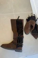 Botas de caña alta confeccionadas en cuero nobuk color chocolate, con detalle de flecos laterales en color negro, suela plana y cierre lateral.