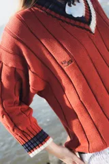 Buzo tejido de punto con escote en V, manga larga y diseño holgado. Presenta un patrón de rayas verticales en relieve y detalles de franjas en contraste en el cuello, puños y ruedo. Incluye un pequeño bordado en el pecho.