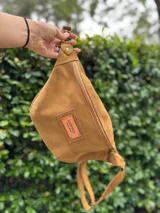 Riñonera de gamuza color tostado con cierre frontal y etiqueta de cuero con logo de la marca Antonia Wool Wear. Cuenta con correa ajustable y herrajes metálicos.