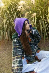 Buzo tipo canguro oversize confeccionado en paño de lana con estampado a cuadros en tonos verdes, amarillos y negros. Presenta una capucha amplia de color violeta, cuello con cierre y detalles en cuero sintético marrón.