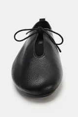 Zapato plano tipo bailarina confeccionado en piel con textura granulada. Presenta cierre mediante lazo en la parte delantera, punta redonda y tirador en la parte trasera.