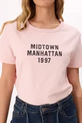 Remera de algodón color rosado con cuello redondo y mangas cortas. Presenta un estampado frontal en color negro con el texto 'MIDTOWN MANHATTAN 1997' en tipografía estilo varsity.