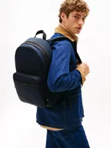 Mochila de poliéster color azul marino con ribetes negros. Presenta un diseño liso, asa superior, correas ajustables y acolchadas, y un panel trasero de malla acolchado. Cuenta con un compartimento principal amplio, funda para portátil incorporada, bolsillo interior con cremallera y bolsillo exterior con cremallera, donde se ubica el logo metálico de Tommy Hilfiger.