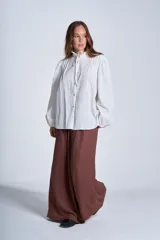 Camisa de color azul con cuello alto decorado con volados y lazo de ajuste. Presenta tablas decorativas en los hombros, cierre frontal con botones de nácar y mangas largas con puños elásticos que generan un efecto abullonado.
