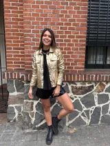 Chaqueta dorada metalizada con cuello camisero, cierre frontal con botones y bolsillos de parche en el pecho.