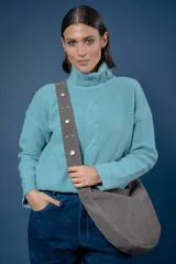 Bolso tipo bandolera de gamuza color azul, con correa ancha ajustable decorada con tachas metálicas circulares. Presenta un diseño de media luna con cierre superior.