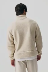 Campera deportiva de tejido sherpa color beige, con cuello elevado, cierre frontal completo, mangas raglán, bolsillo superior con cierre y bolsillos laterales. Presenta terminaciones elásticas en puños y ruedo.