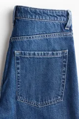 Jeans de tiro alto, pierna ancha y corte recto, confeccionado en denim de algodón rígido de color azul medio. Presenta un detalle de herrajes metálicos en la cintura.