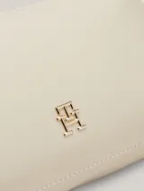 Cartera rectangular tipo bandolera de color blanco roto, con acabado liso y herrajes dorados. Presenta un monograma metálico TH en el centro y una correa ancha ajustable y extraíble.