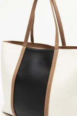 Cartera tipo tote bag en símil cuero, con diseño en bloques de color negro y blanco, y detalles en marrón. Tiene asas largas para llevar al hombro y un cierre magnético.