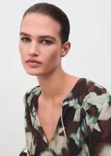 Blusa de gasa con estampado floral en tonos verdes y marrones, cuello escotado con cierre de lazo y mangas largas con puños abotonados.