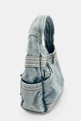 Bolso de mano tipo hobo confeccionado en tejido denim con diseño inspirado en un jean, incluyendo detalles de bolsillos exteriores, costuras reforzadas y trabillas. Cuenta con cierre de botón imantado y asa integrada.