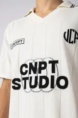 Remera tipo chomba de tejido de punto color blanco, con cuello polo y diseño de rayas verticales sutiles. Presenta estampado gráfico frontal con texto y logo, además de detalles de rayas negras en los hombros.