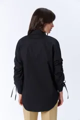 Camisa negra de manga larga con cuello clásico y cierre frontal con botones. Las mangas tienen frunces y lazos para ajustar el largo.