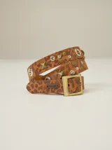 Cinto de cuero con estampado animal print de leopardo, decorado con tachas metálicas, remaches y ojales. Cuenta con una hebilla cuadrada de metal y una placa metálica con el logo de la marca.