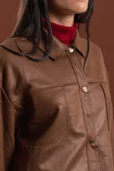Chaqueta de cuero sintético color marrón, con diseño de corte recto, cuello camisero, cierre frontal con botones metálicos y bolsillos frontales tipo parche.