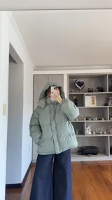 Campera puffer de corte holgado en color beige, con diseño acolchado, cuello alto y capucha desmontable. Cuenta con cierre frontal y botones a presión, además de puños elásticos.