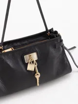 Bandolera de cuero vacuno color negro con efecto brillante, herraje metálico tipo candado y dijes de llave colgantes en tono dorado. Cuenta con correa fina ajustable y tiras laterales decorativas.