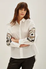 Camisa blanca de algodón con mangas largas, destacada por bordados decorativos en color negro y plateado en las mangas.