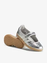 Zapatillas Adidas Samba Jane W de color plateado, con diseño híbrido entre zapatilla y Mary Jane, confeccionadas en piel y material sintético. Presentan tres bandas laterales blancas, tira ajustable con cierre de velcro en el empeine, y suela de goma de tipo caramelo.