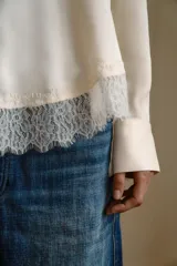 Camisa de satén color crema con cuello solapa, manga larga y cierre frontal con botones ocultos. Presenta un detalle de encaje en el bajo asimétrico.