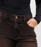 Pantalón de jean corte flare con lavado sobretinta en tono marrón oscuro. Presenta cintura media con trabillas, cierre frontal con cremallera y botón, cinco bolsillos funcionales y ruedo deshilachado.