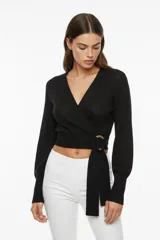 Sweater negro de punto con diseño cruzado tipo wrap, cuello en V y mangas largas con puños acanalados. Presenta un detalle de argolla metálica dorada en la cintura donde se anuda la prenda, con un corte corto al cuerpo.